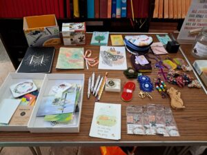 Table of handmade items on a wooden surface: notebooks, greeting cards, keychains, beaded bracelets, crosses, and small crafts for sale.
